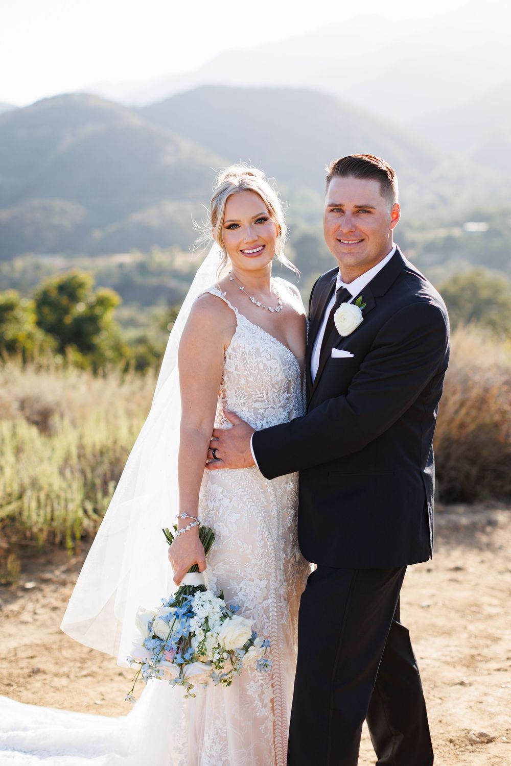A wedding couple poses together in a scenic desert mountain landscape with a beautiful bouquet of blue flowers.
