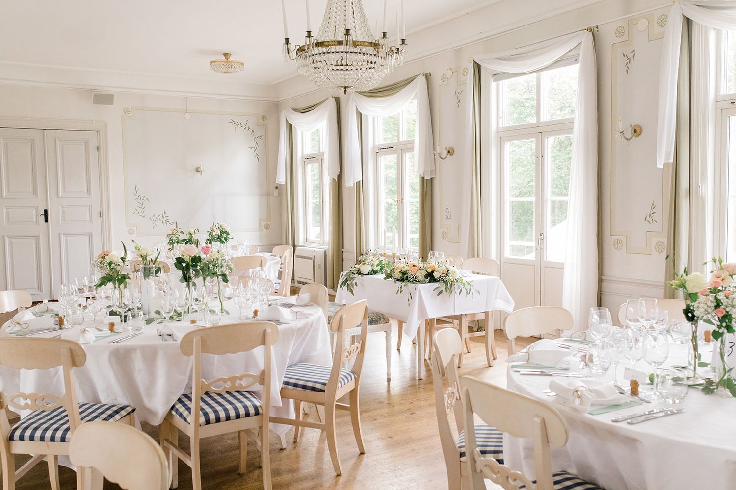 Elegant white wedding reception hall with round tables, chandeliers and large windows creating a bright, airy atmosphere.