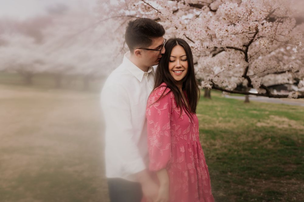 A couple embraces under blooming cherry blossom trees in a dreamy spring setting with soft pink tones.