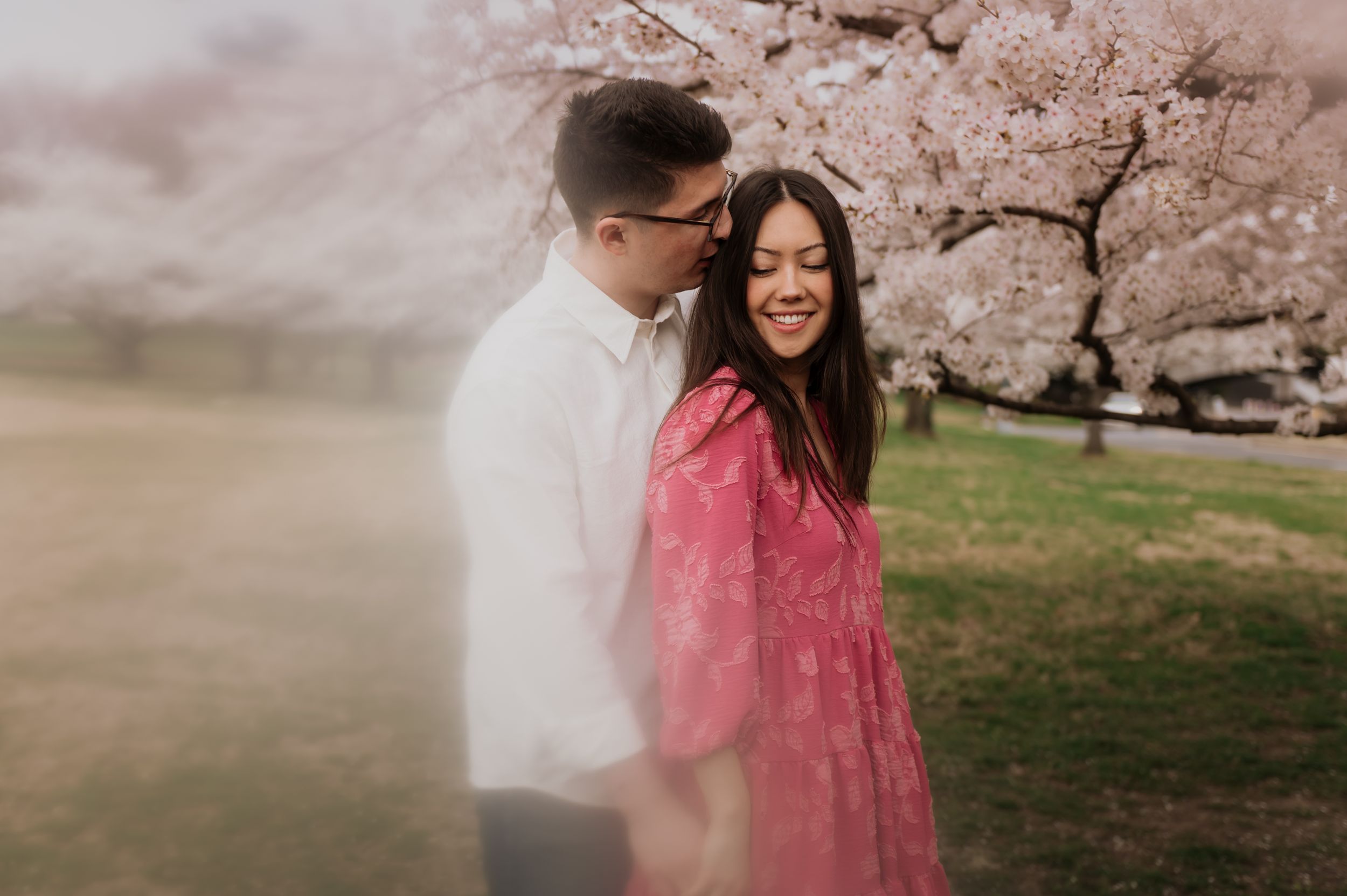 A couple embraces under blooming cherry blossom trees in a dreamy spring setting with soft pink tones.