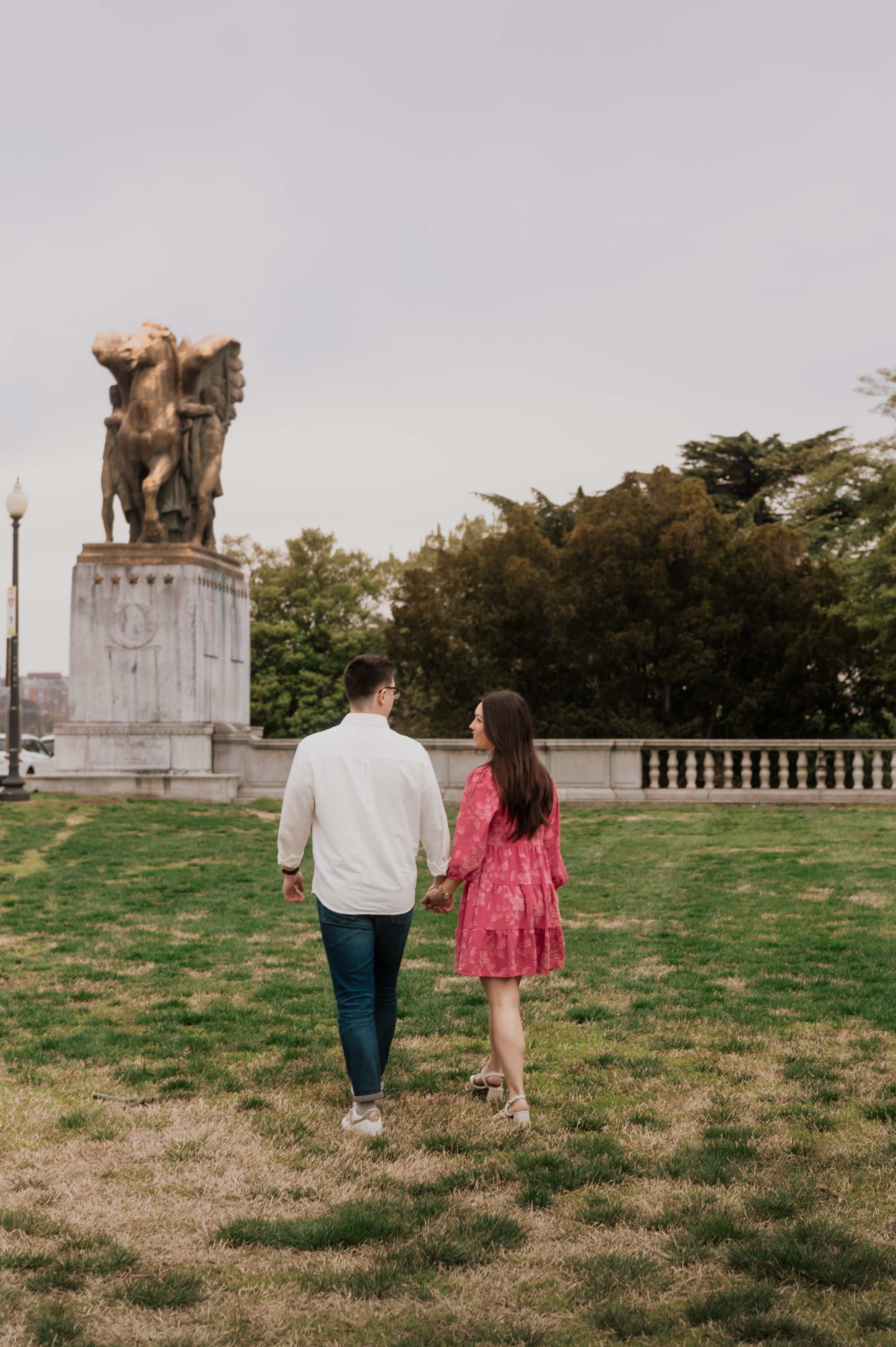 A couple walks hand in hand on a grassy lawn in front of a large equestrian statue monument.