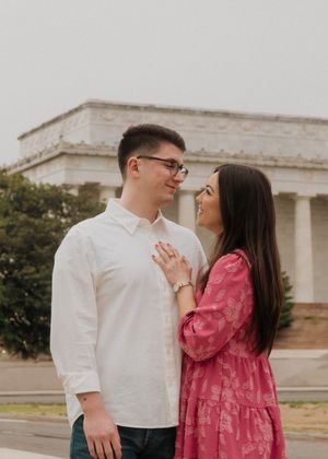 A couple shares a romantic moment wearing white and pink outfits in front of a classic stone building during a photo session.
