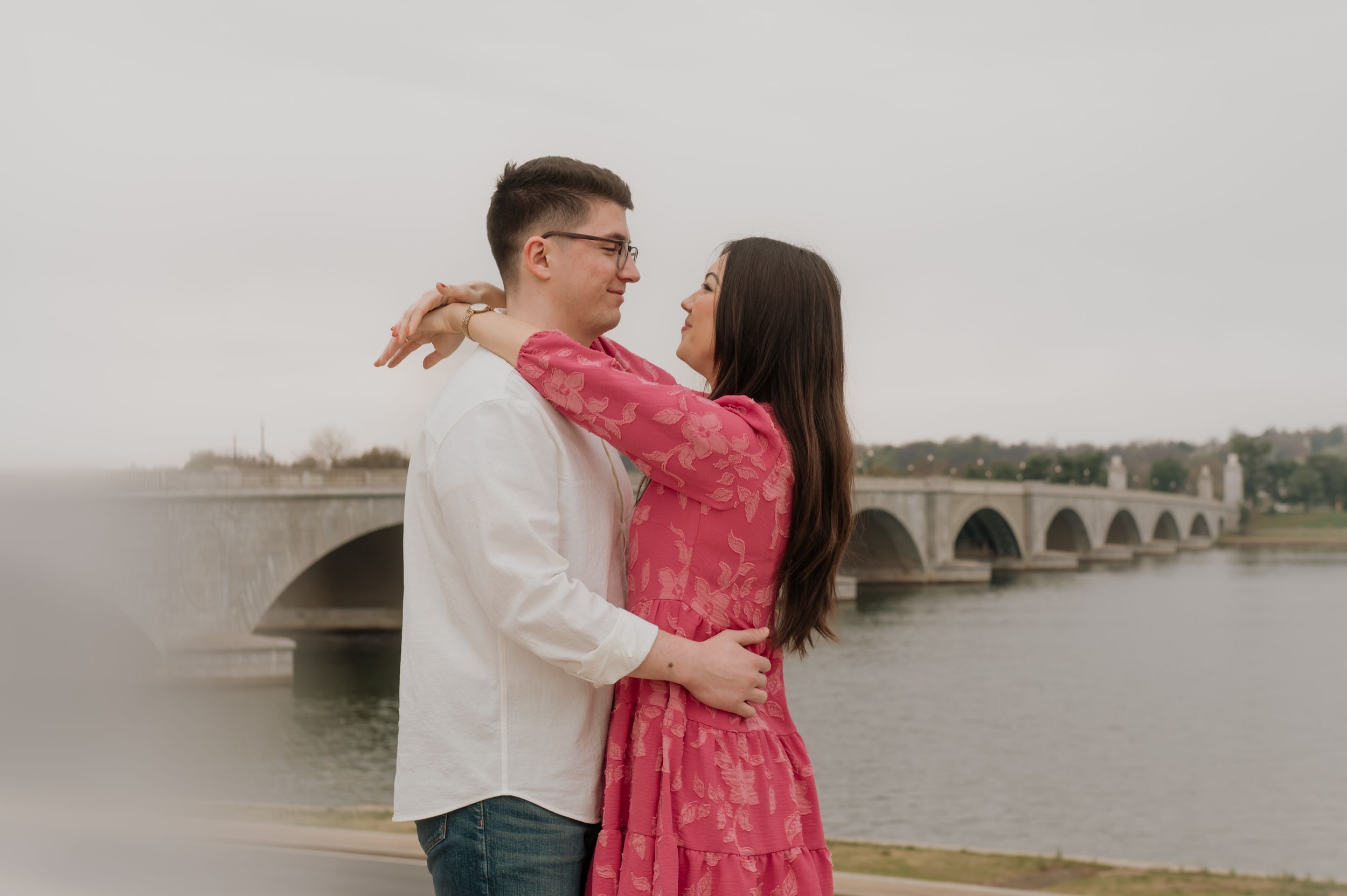 Couple embraces near a stone bridge over water with person in pink dress and white shirt.