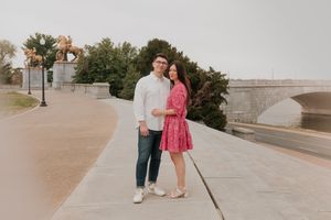A couple stands together on a concrete walkway near palm trees and a bridge during a sunset photoshoot.