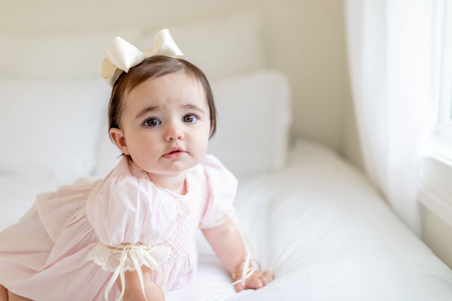 Baby wearing pink dress sits on white bed near window with soft natural lighting.