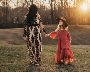 A mom and a daughter in dresses walking holding hands on a hill in front of the sunset