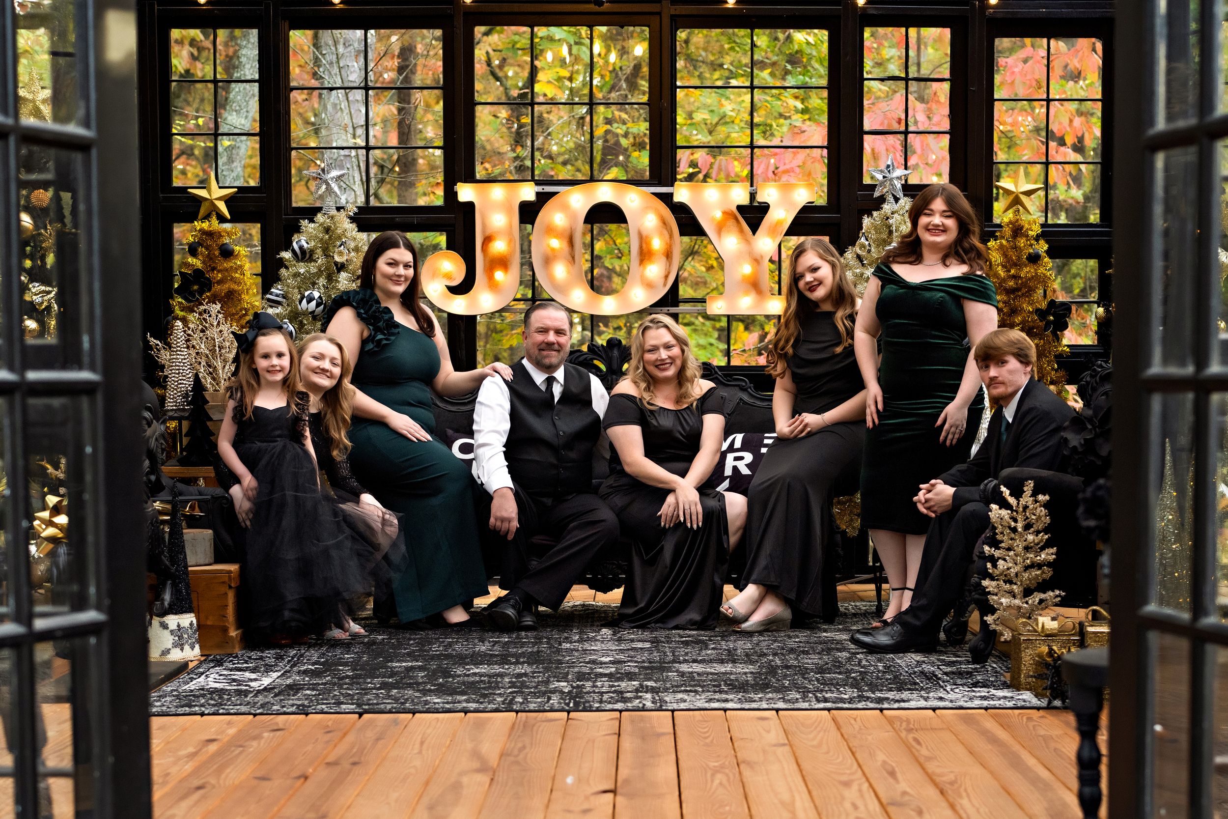 A large family sitting on a couch in a black and gold greenhouse that is decorated for Christmas with the large word JOY in the background