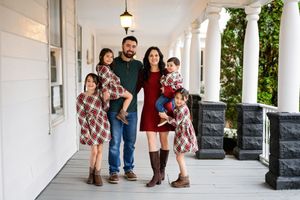 A family of 5 dressed for Christmas standing on a large white porch of a historic house