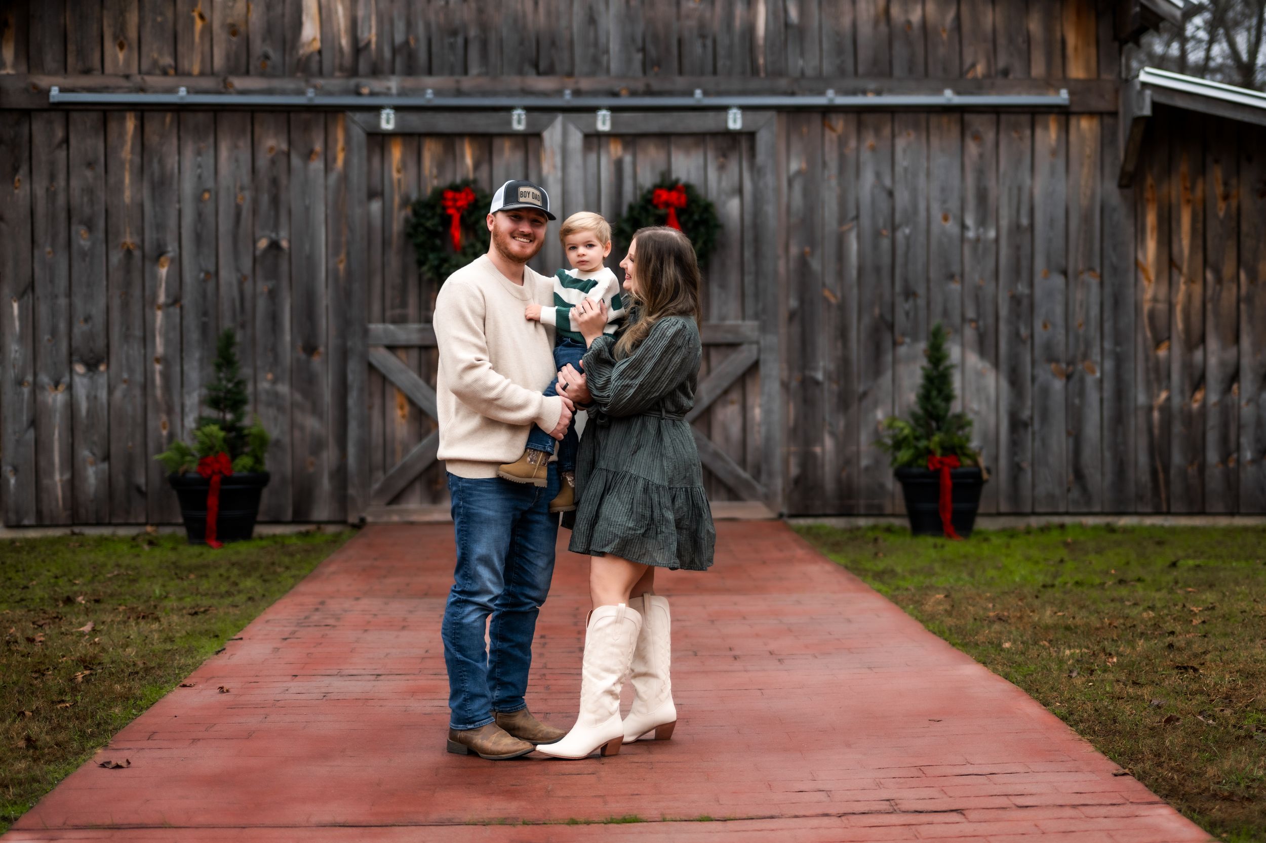 A family of three standing in front of a wooden brown barn decorated for Christmas