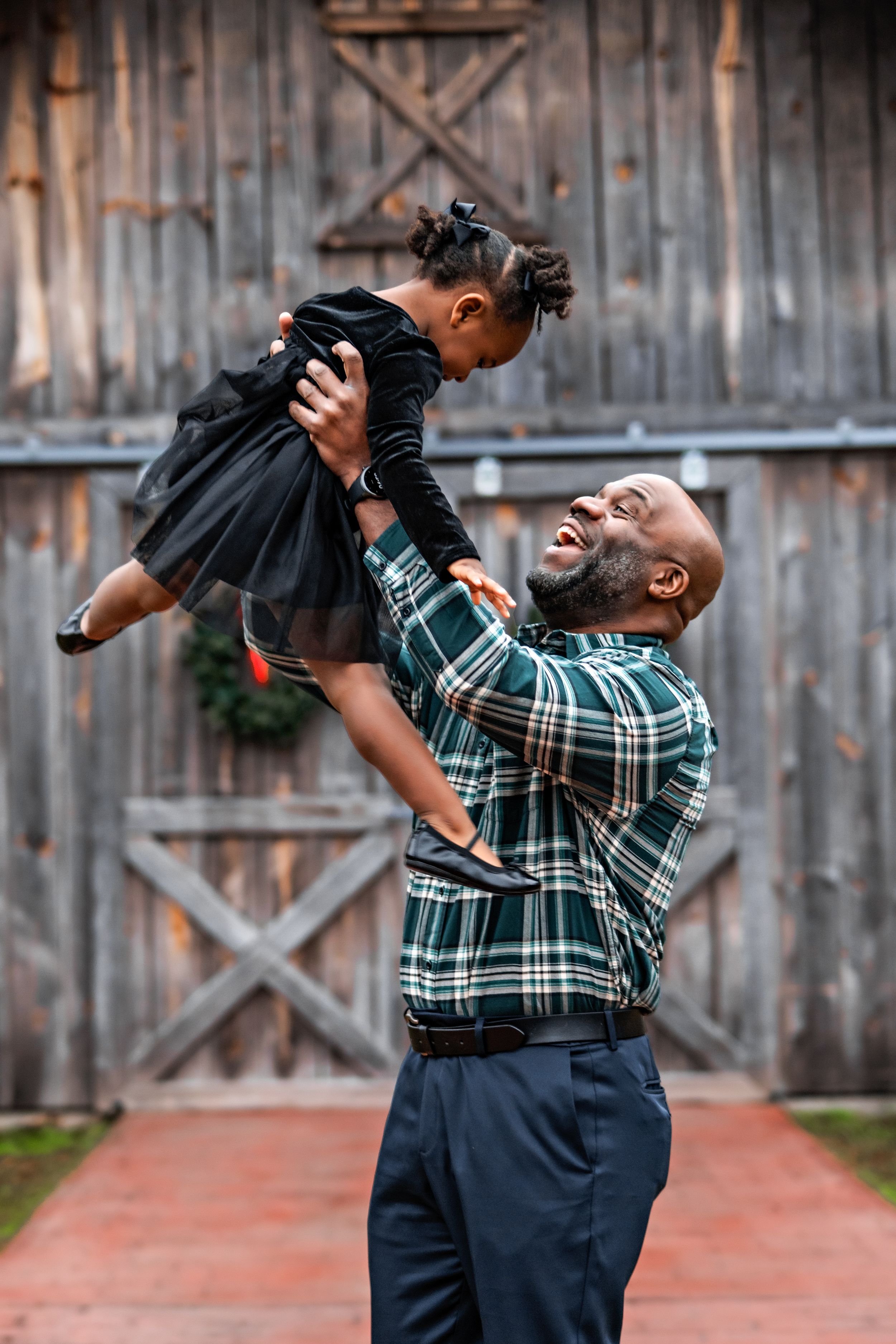 a dad lifting his toddler daughter into the air in front of a brown wooden barn