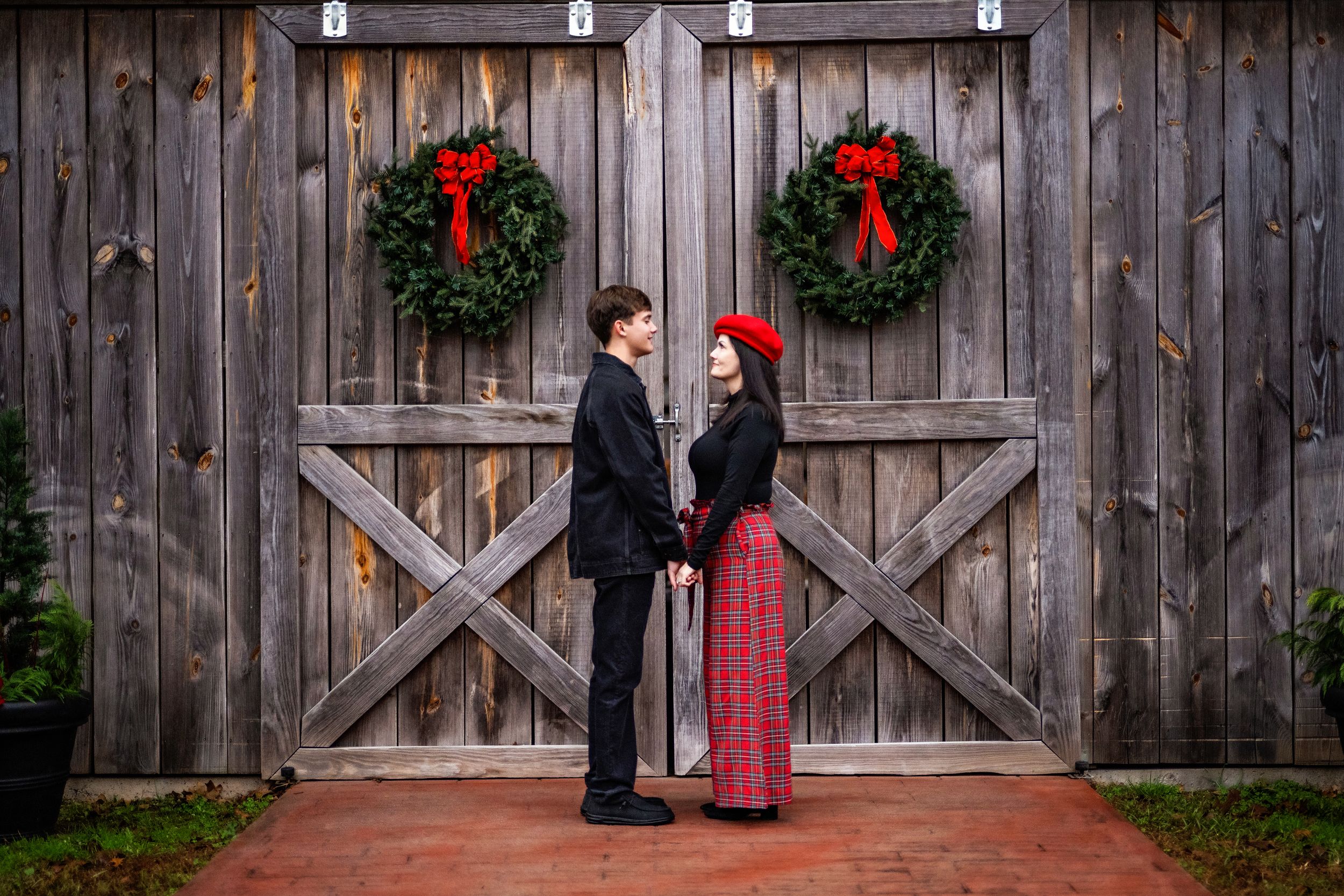 A mom and son standing holding hands facing each other in front of a large brown wooden barn that is decorated for Christmas