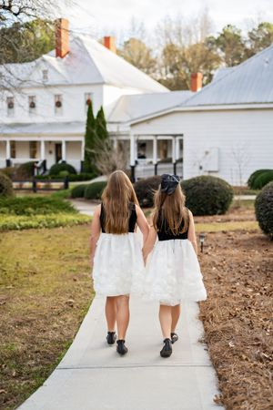 Two sisters walking hand in hand beside each other down a path towards a white historic house