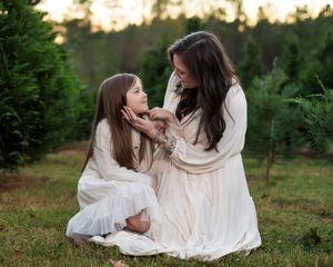 A mom and daughter sitting on the ground in a Christmas tree farm. The mom is holding the daughter's face