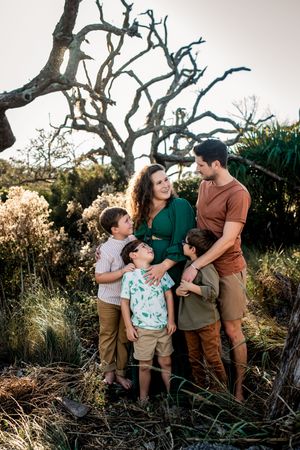 A family of 4 standing on a beach in front of greenery and driftwood trees