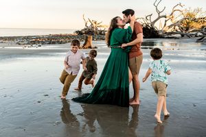 A husband and wife hugging each other on a beach as their 3 children run around them playing