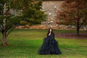 A lady in a black formal dress standing on the grass next to a tree in front of a stone building