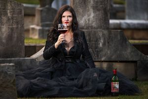 A lady in a black formal dress sitting in a cemetery holding a glass of wine in front of her face.