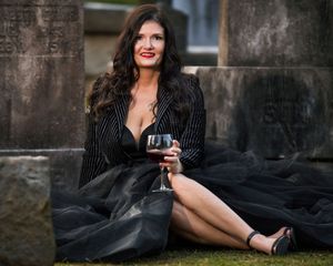 A lady in a black formal dress sitting in a cemetery holding a glass of wine