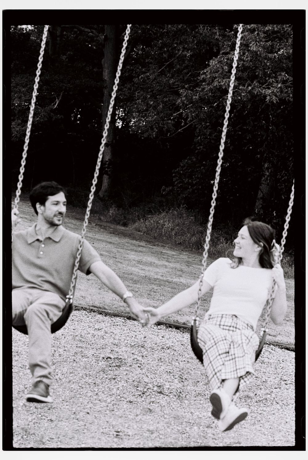 Candid scene of couple swinging together on a park swing set while they hold hands and laugh and smile at each other in vacationland.