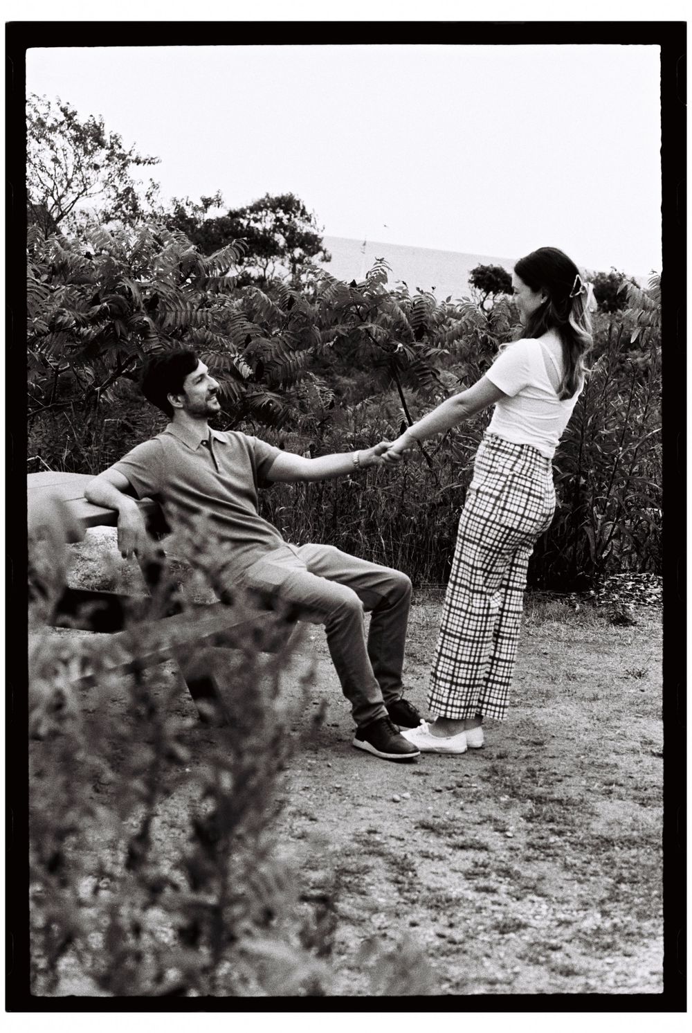 Candid black and white film photo of couple as he sits and she stands holding his hand pulling him up towards her in Cape Elizabeth, Maine.