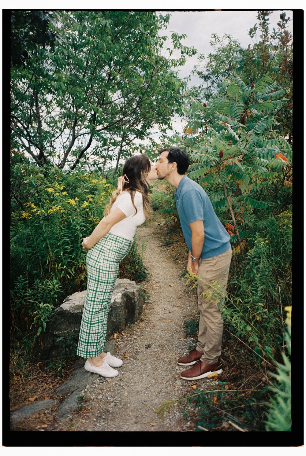 Couple stands on either side of a park pathway leaning into kiss each other surrounded by lush greenery on 35mm color film in southern Maine