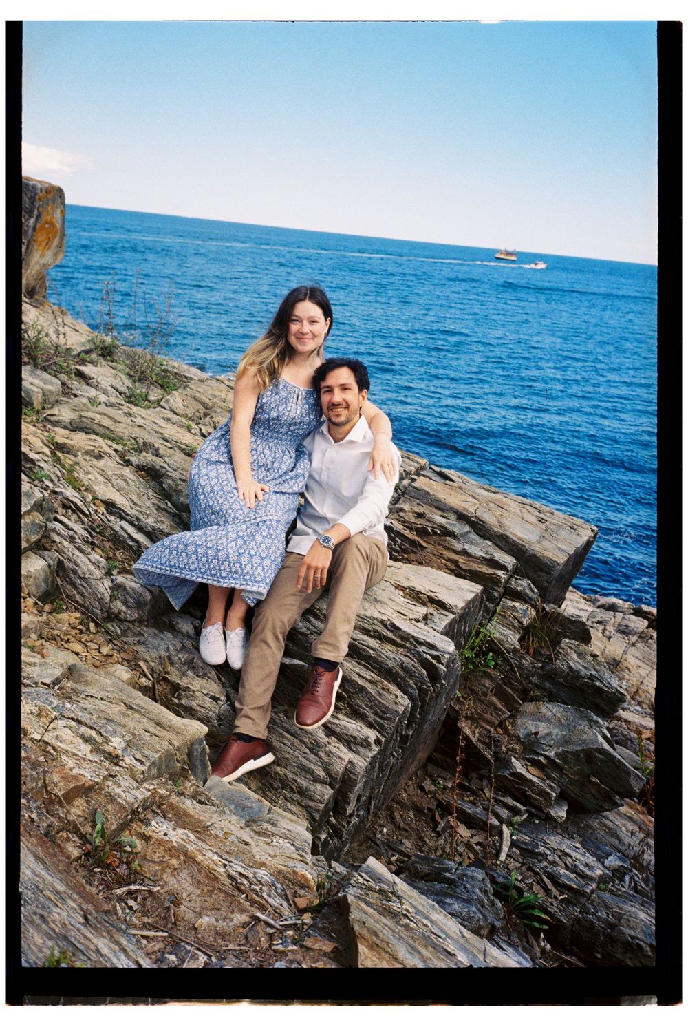 Portrait of a couple on a late summer day as they sit atop big rocks in front of blue skies and blue sea smiling together in Cape Elizabeth.