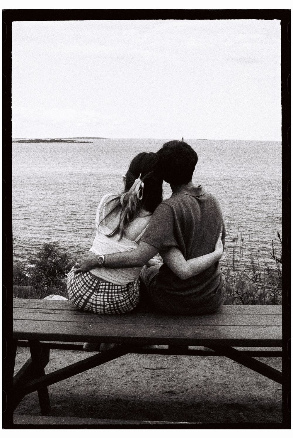 Black and white 35mm film photograph of couple sitting atop a picnic table at the park looking out at the sea, arms wrapped around eachother