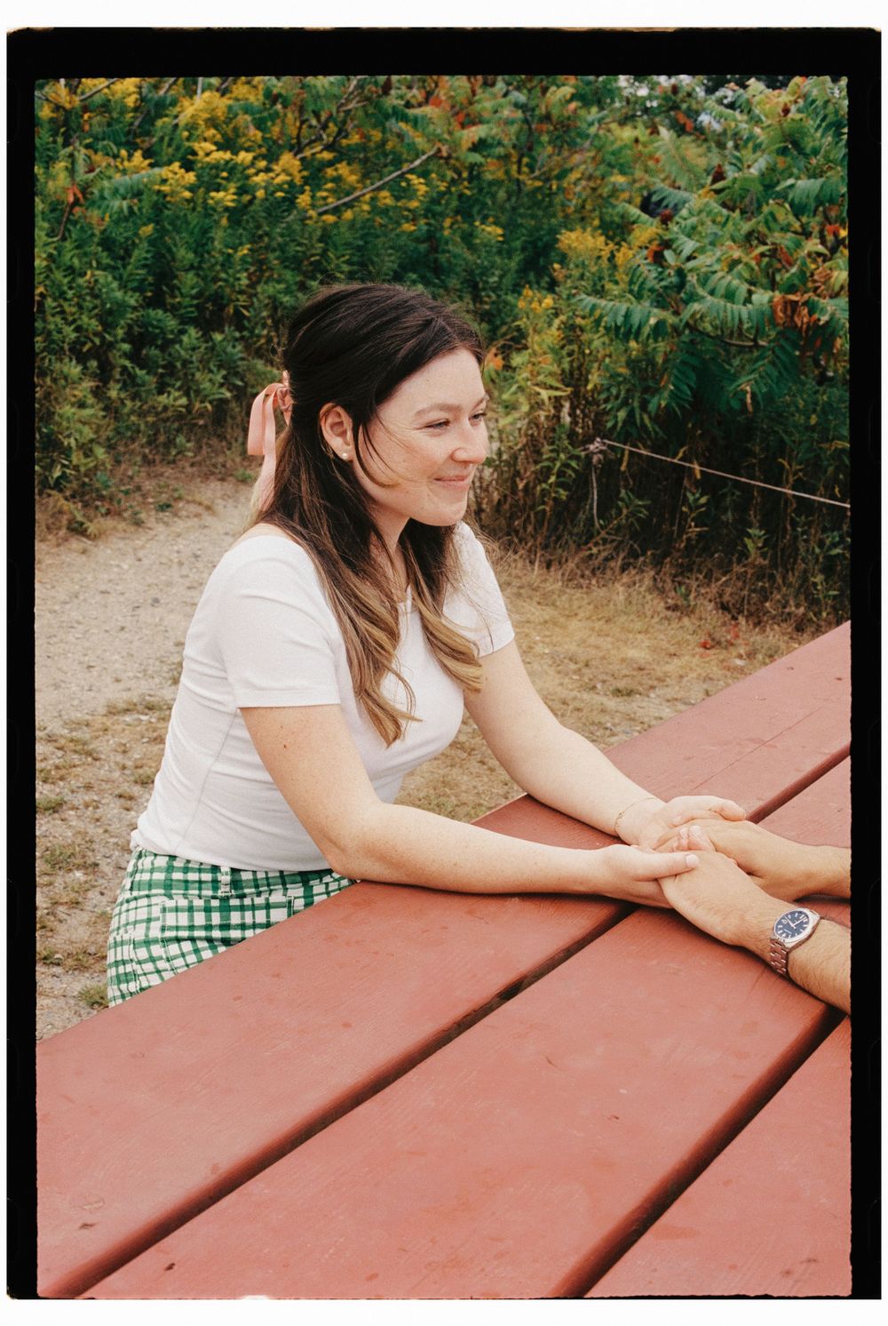 Portrait of her looking at him as they hold hands across a red picnic table with wildflowers growing the background in southern Maine.
