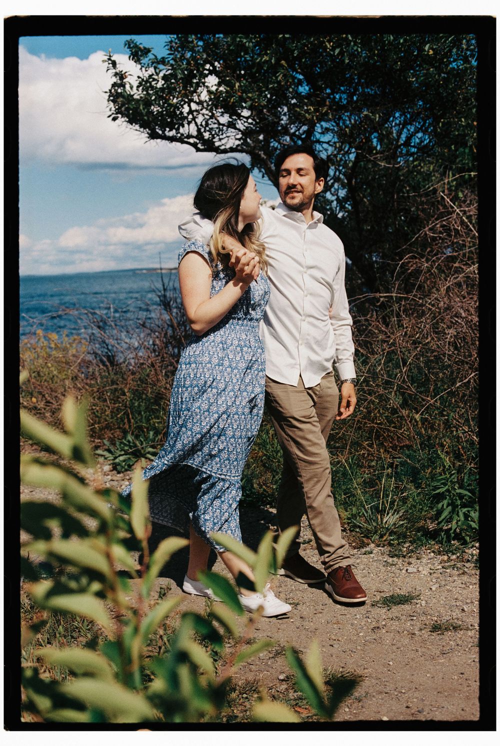 He has his arm around her shoulder as they walk together on a woodland path with the blue ocean and blue skies and clouds behind them.