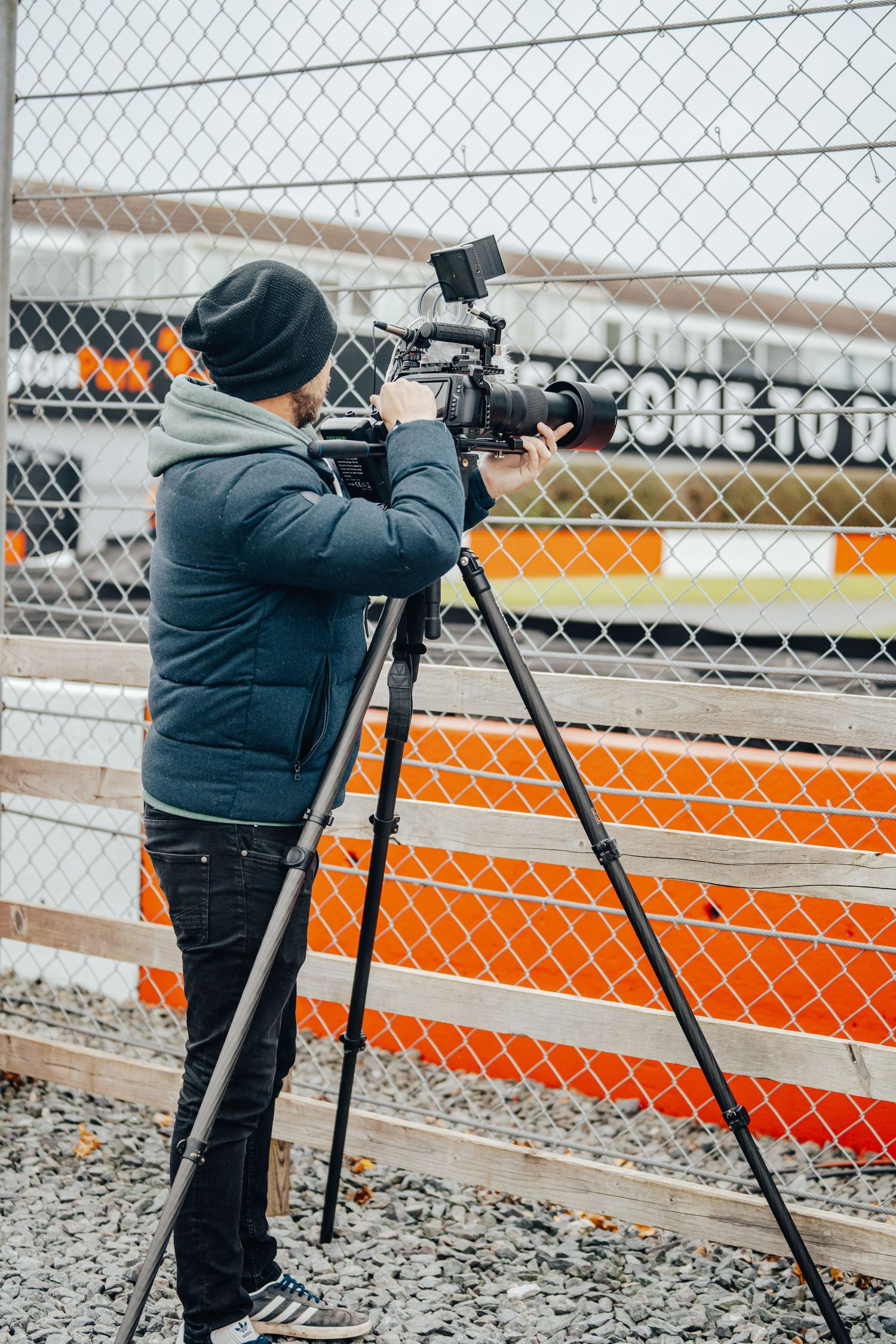 Camera operator films through chain link fence with tripod mounted equipment.