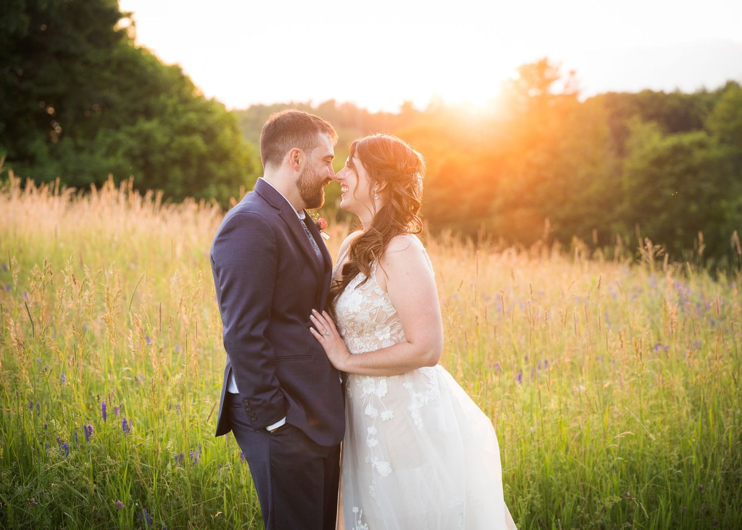 A couple in wedding attire embraces in a sunlit meadow during golden hour as the sun sets behind them.