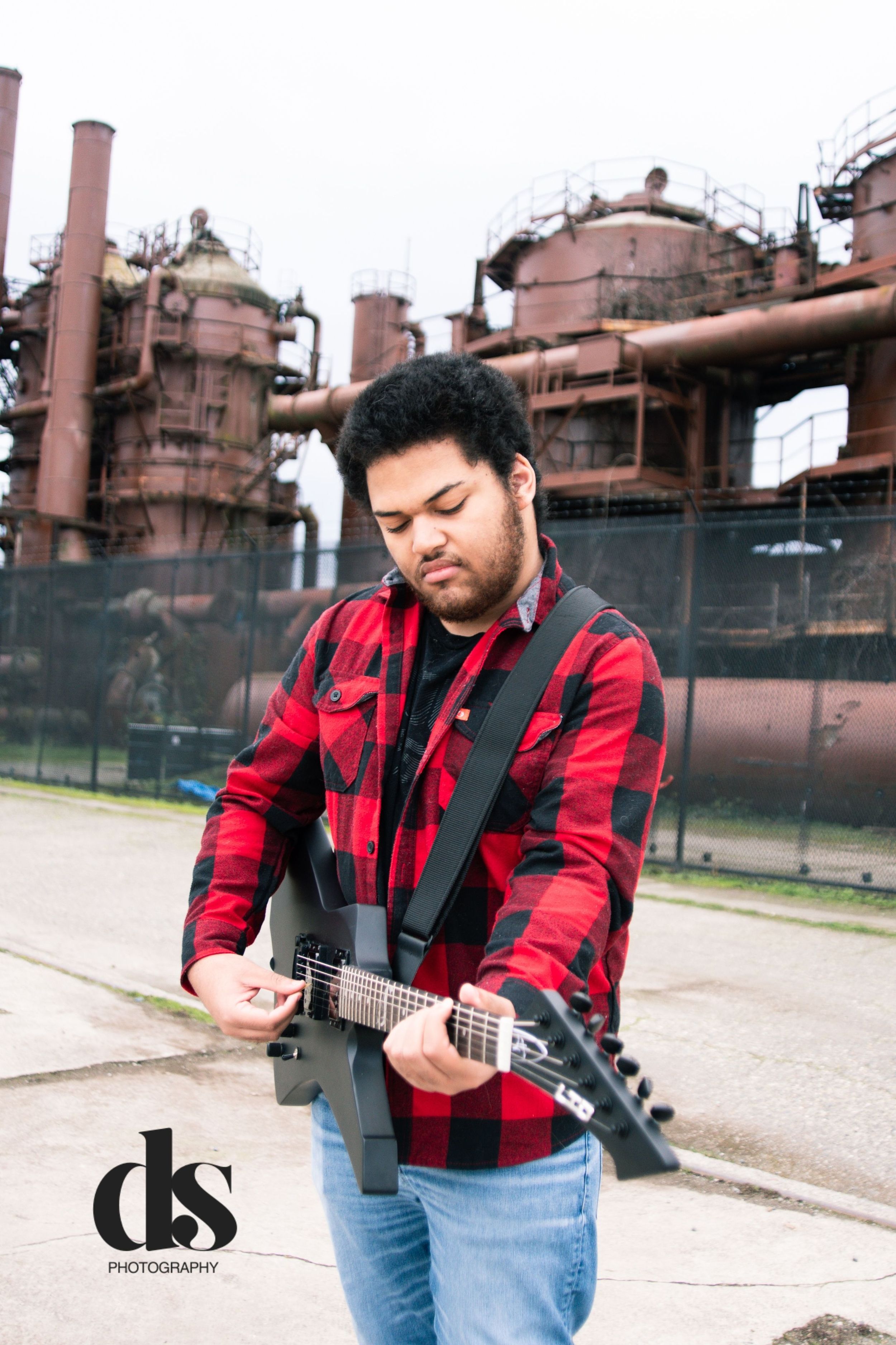 Musician in red plaid shirt plays bass guitar in front of industrial buildings during outdoor photo session.