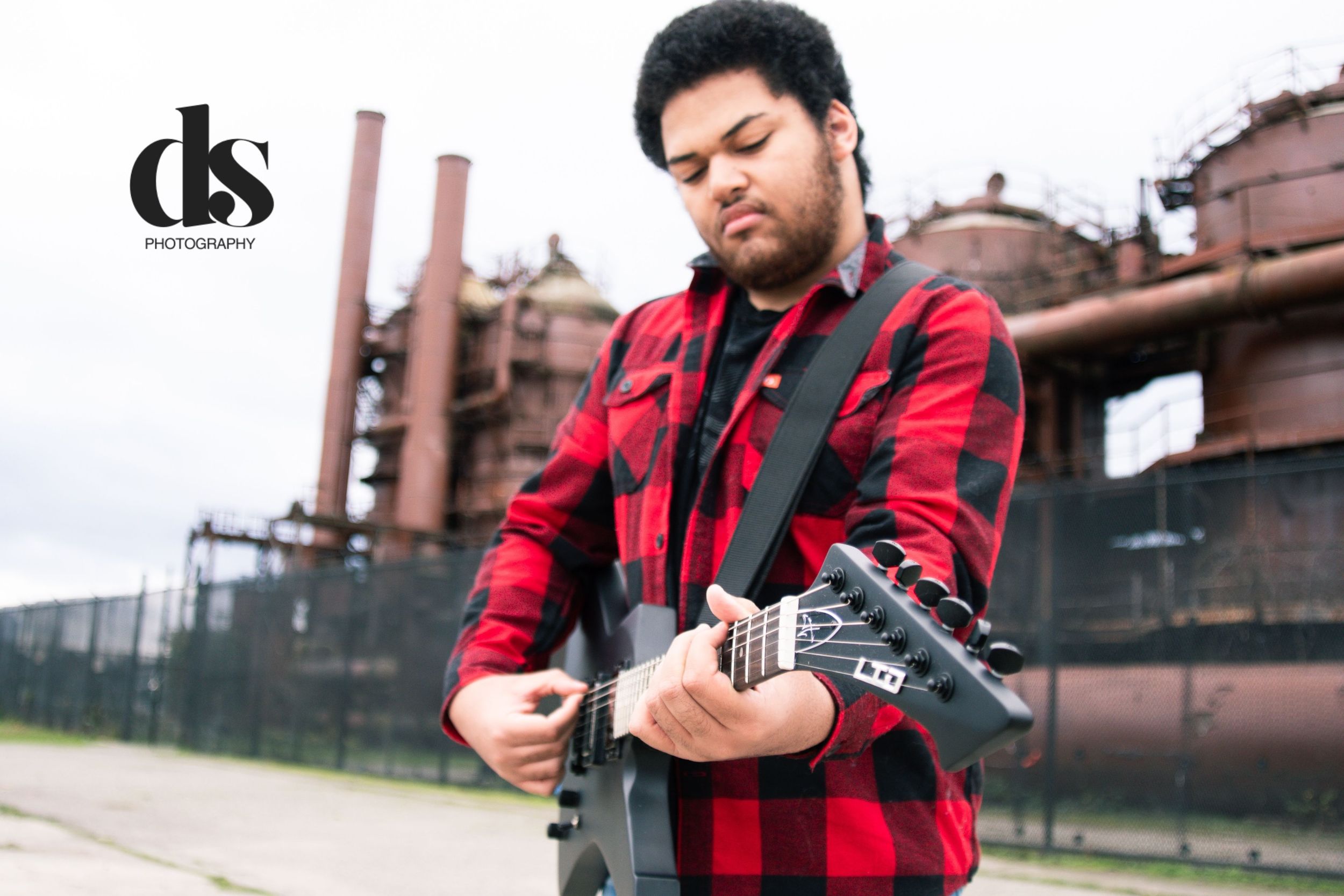 Musician in red plaid flannel shirt plays electric guitar in front of industrial factory backdrop.