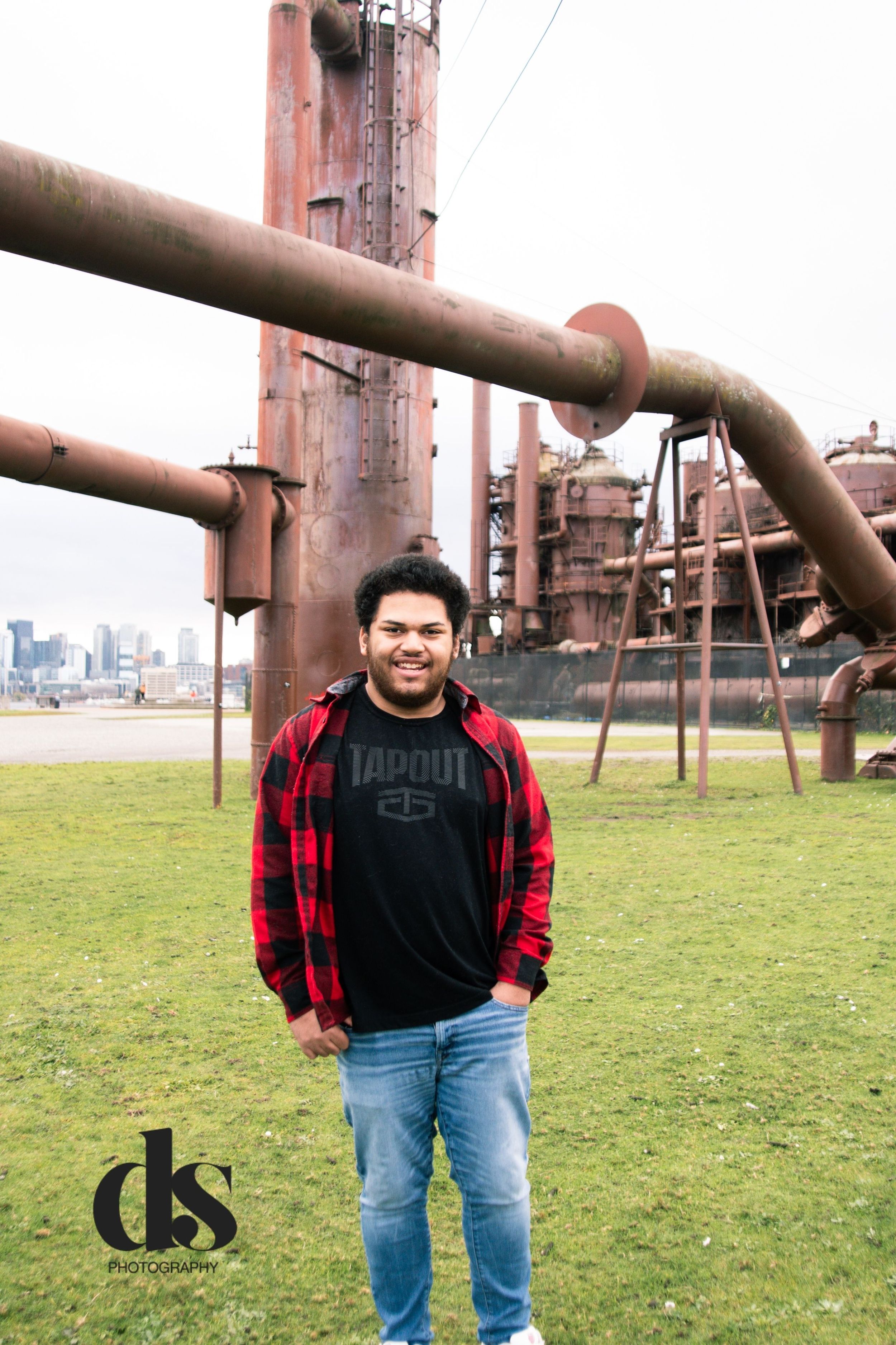 Person in red plaid flannel and jeans poses in front of industrial pipes and equipment at an outdoor location.