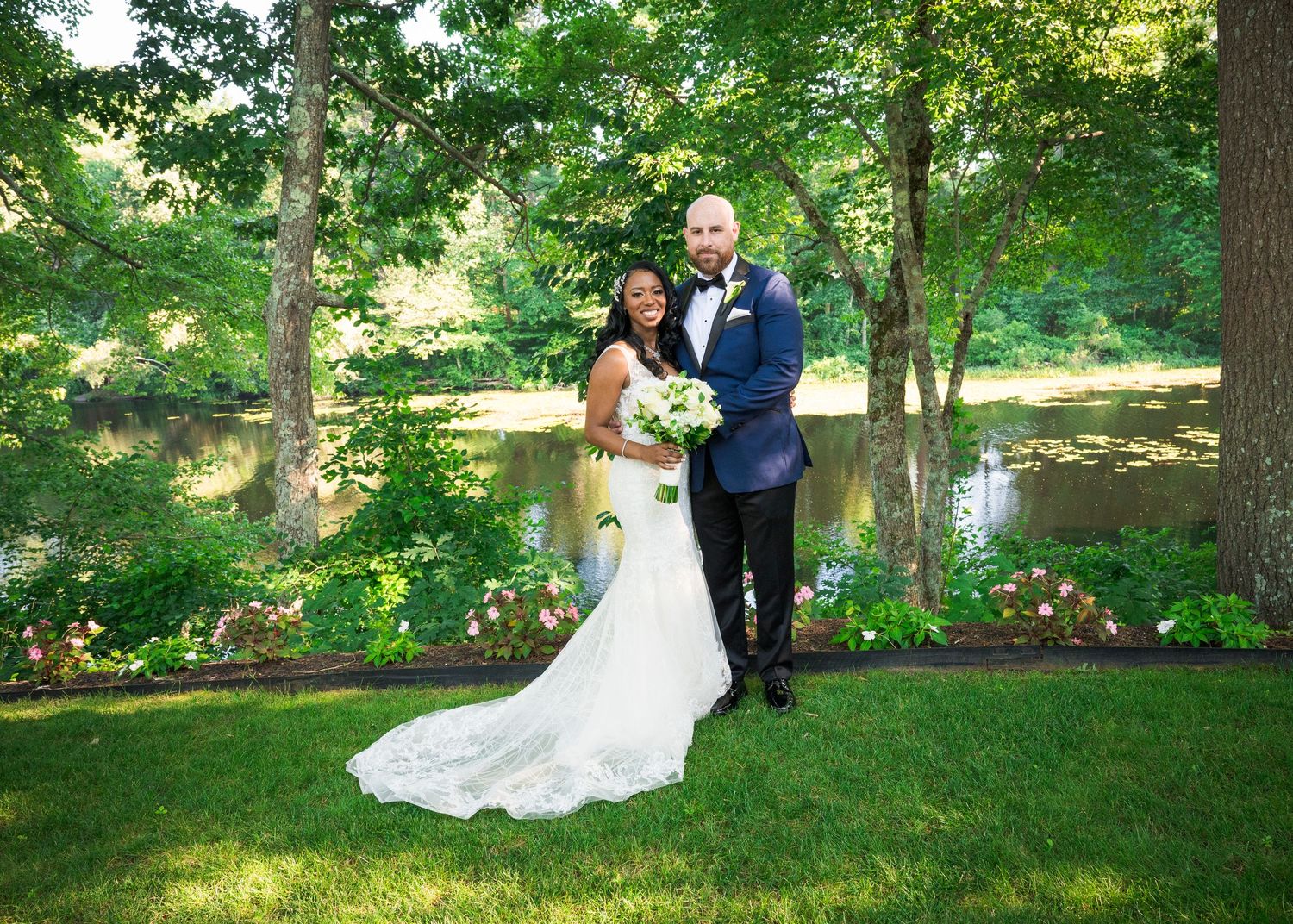 A couple in wedding attire poses together in a lush green garden setting with sunlight filtering through trees.