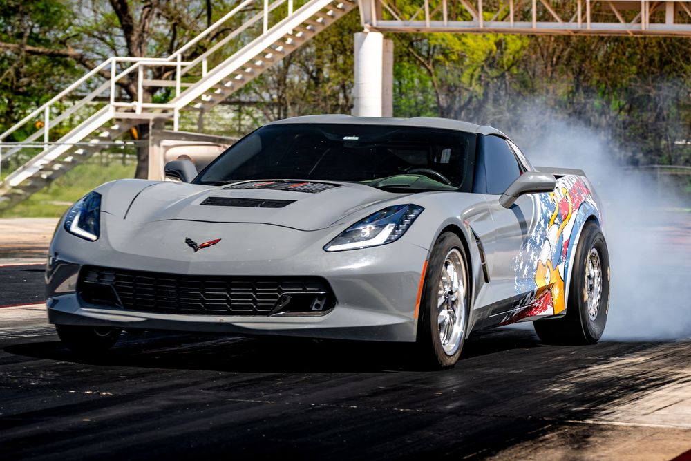 A silver Chevrolet Corvette performs a burnout on a track, creating dramatic tire smoke during a racing event.