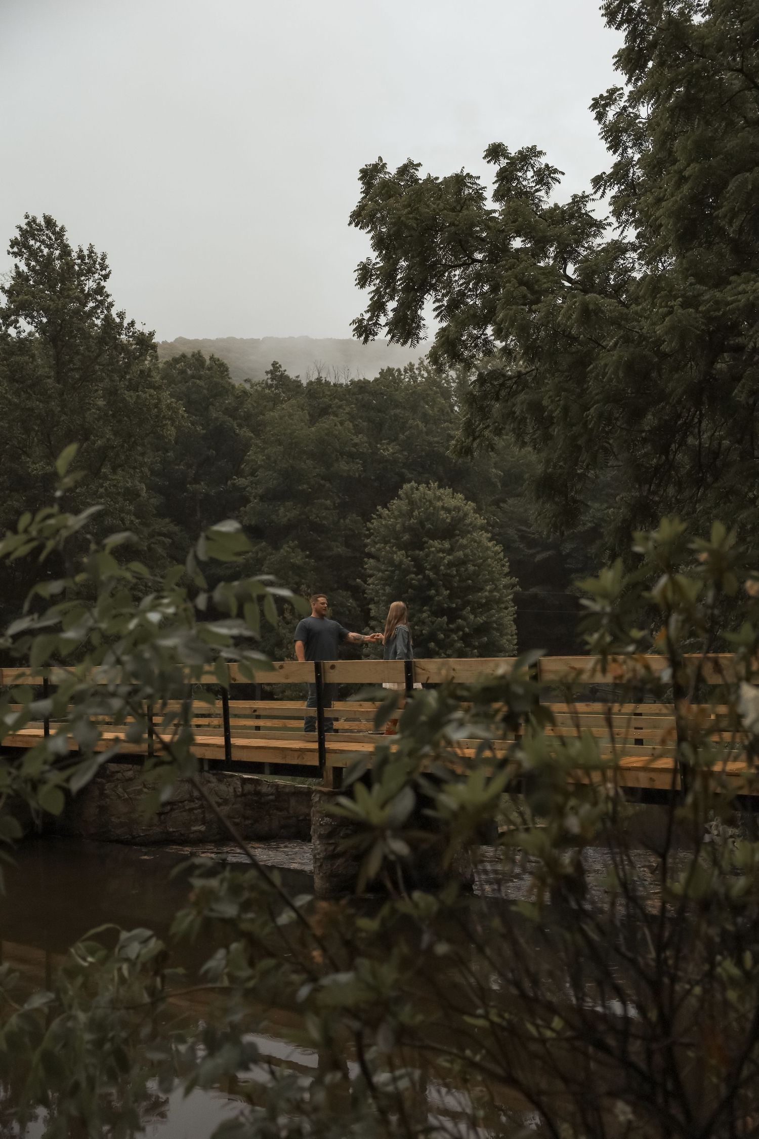 Two people stand on a wooden bridge surrounded by lush green trees on a misty, overcast day.