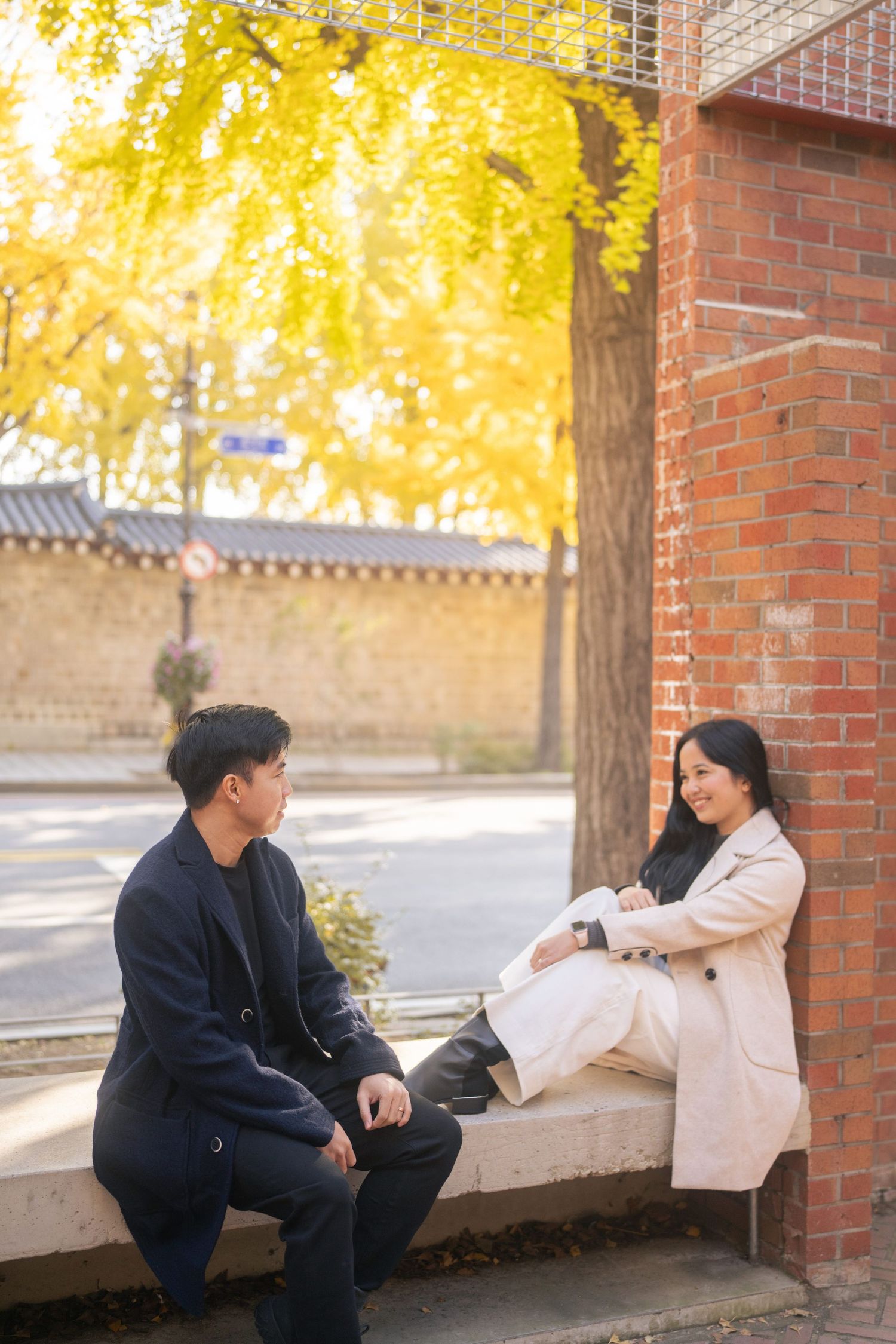 A couple shares a playful moment on a brick ledge surrounded by autumn trees with golden yellow foliage.