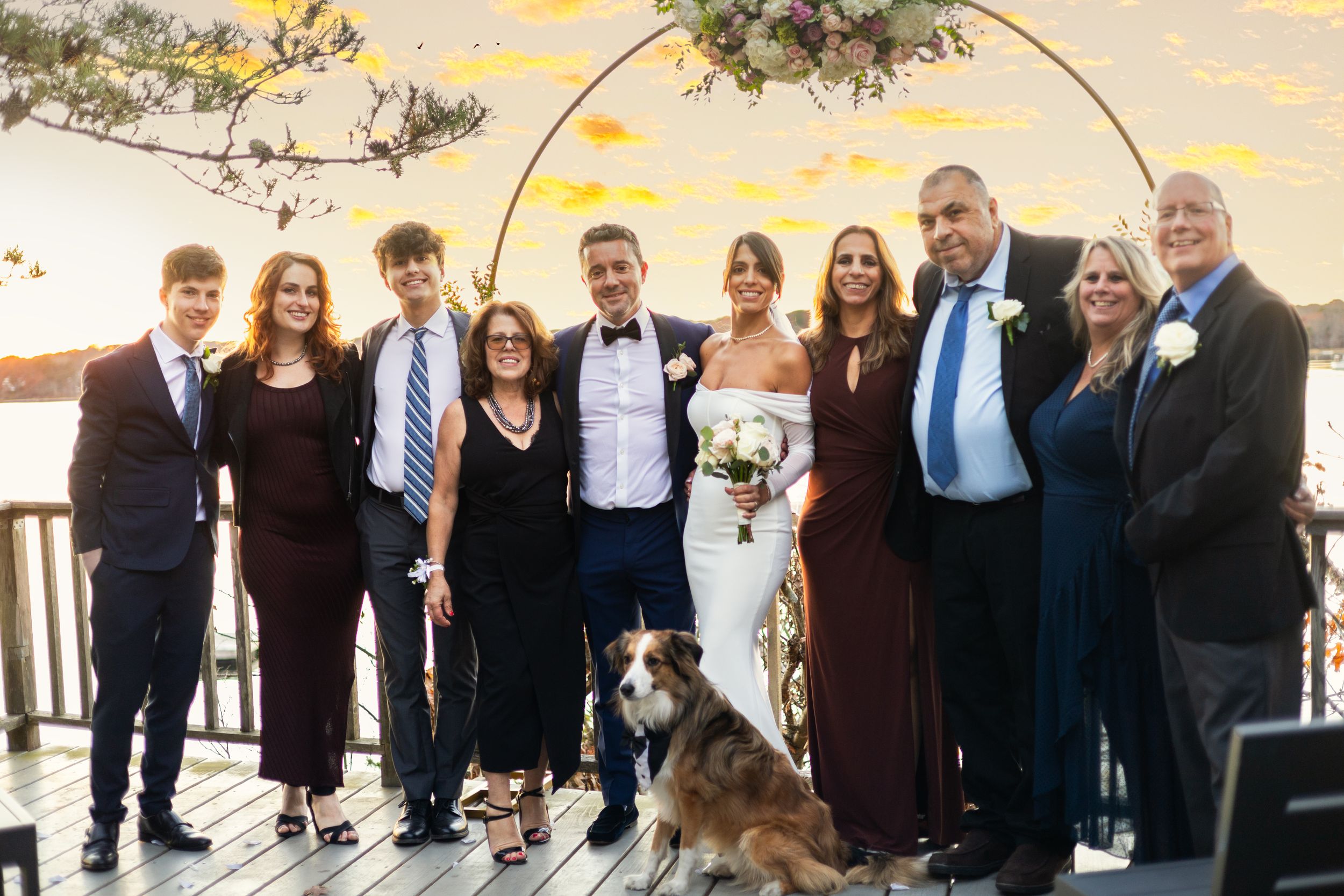 Wedding party group poses at sunset with a dog in front of water view.