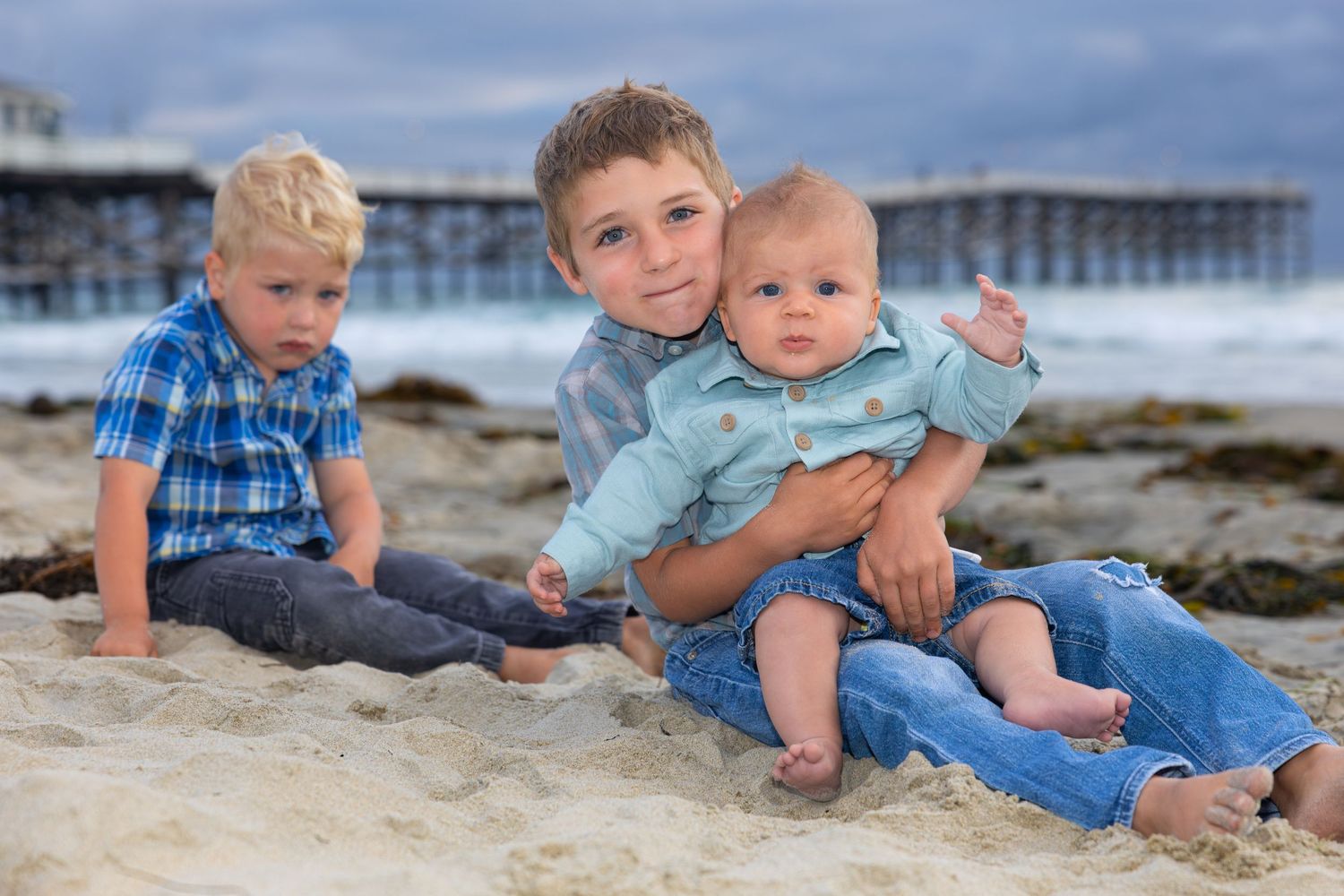 San Diego family photography of three young brothers sitting in the sand at the beach with a wooden pier in the background.