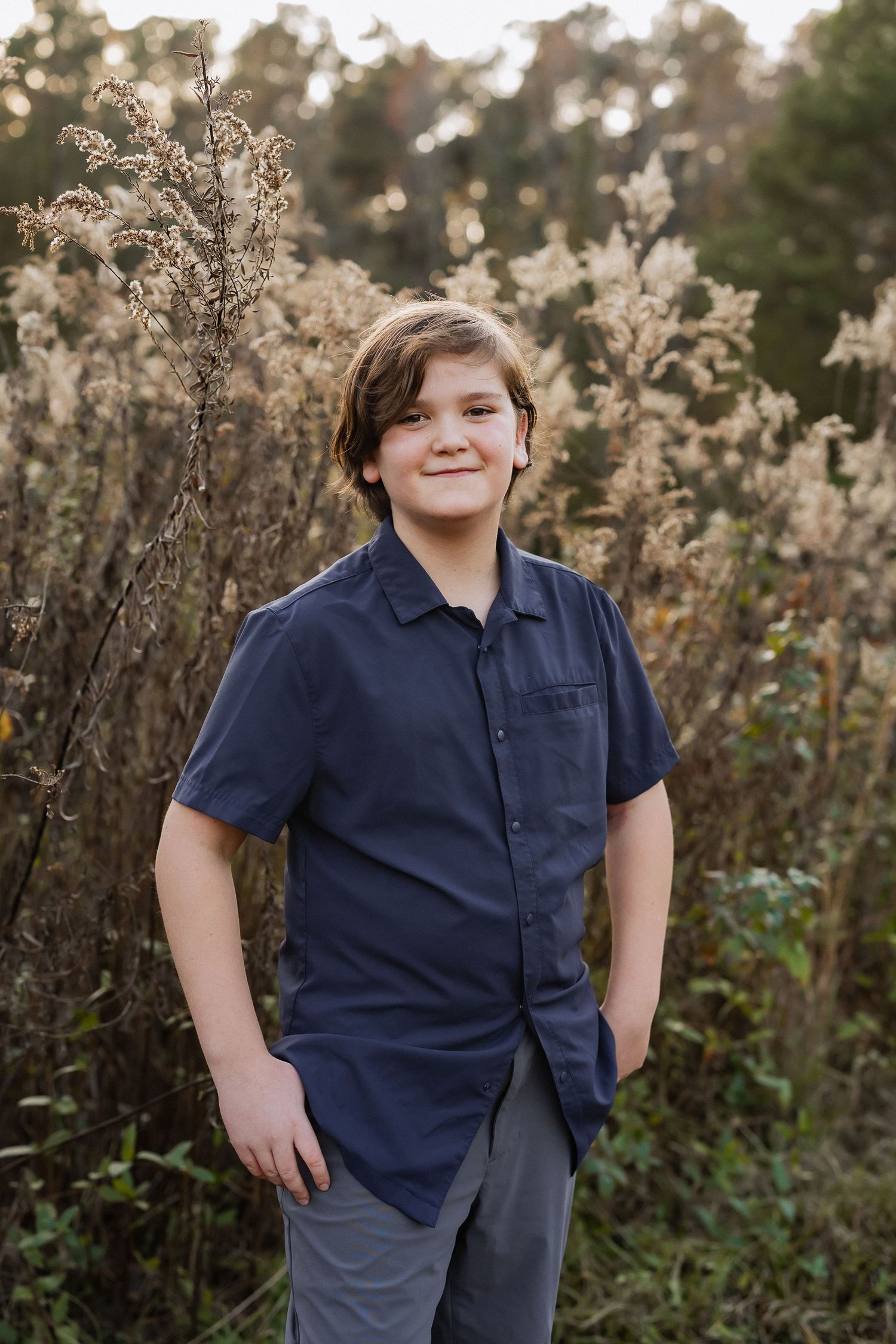 A young person in a navy shirt poses casually against a backdrop of autumn foliage.