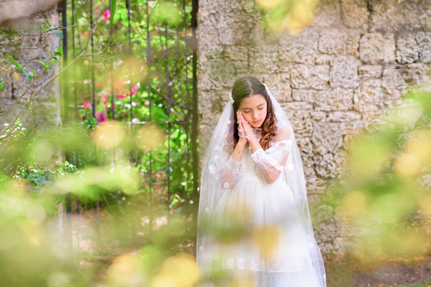 A bride in a white communion dress and veil poses against a stone wall surrounded by soft green foliage.