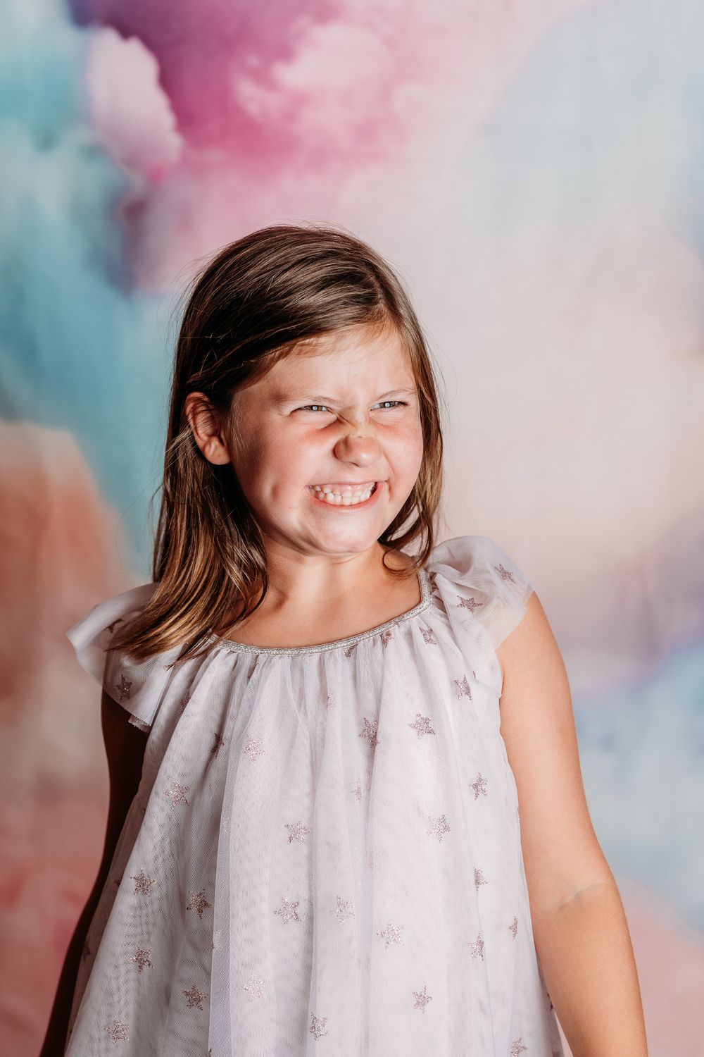 A happy child wearing a light colored dress with star patterns poses against a dreamy pink and blue cloudy background.