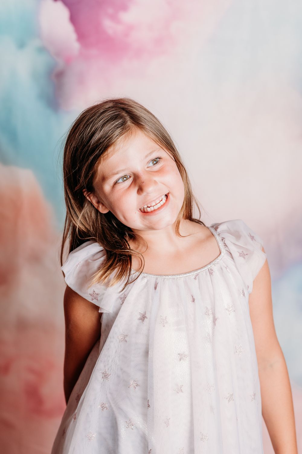 A joyful portrait against a soft pink and blue backdrop showing someone in a white star-patterned dress.