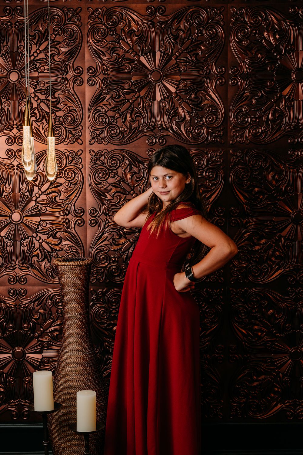 A figure in a flowing red dress poses against an ornate brown metallic wall backdrop with decorative candles nearby.