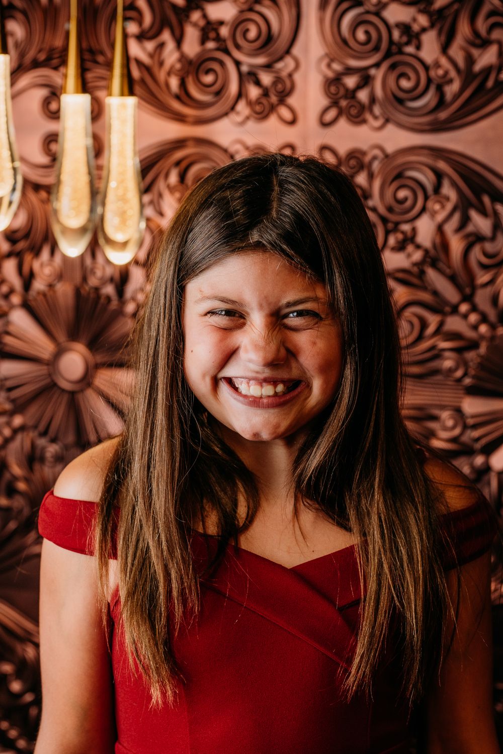 Portrait against ornate copper wall pattern wearing red off-shoulder top and displaying genuine warm smile.