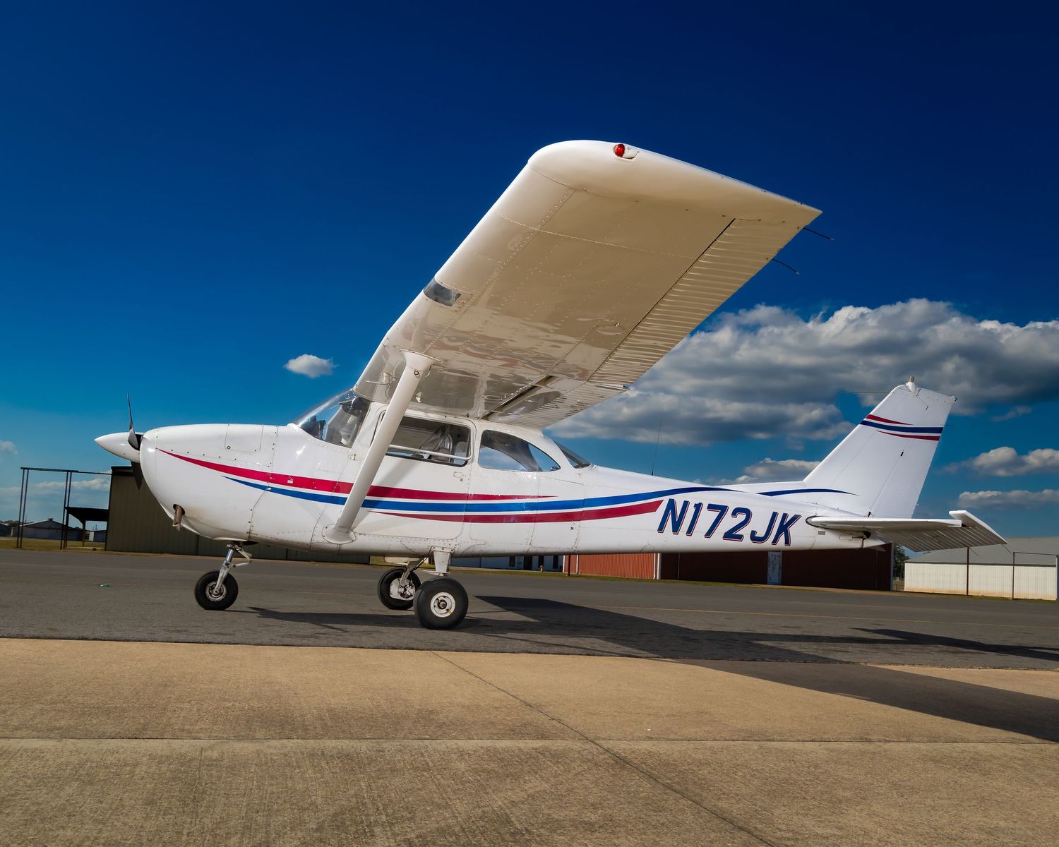 White Cessna aircraft with registration N172JK parked on tarmac against blue sky.