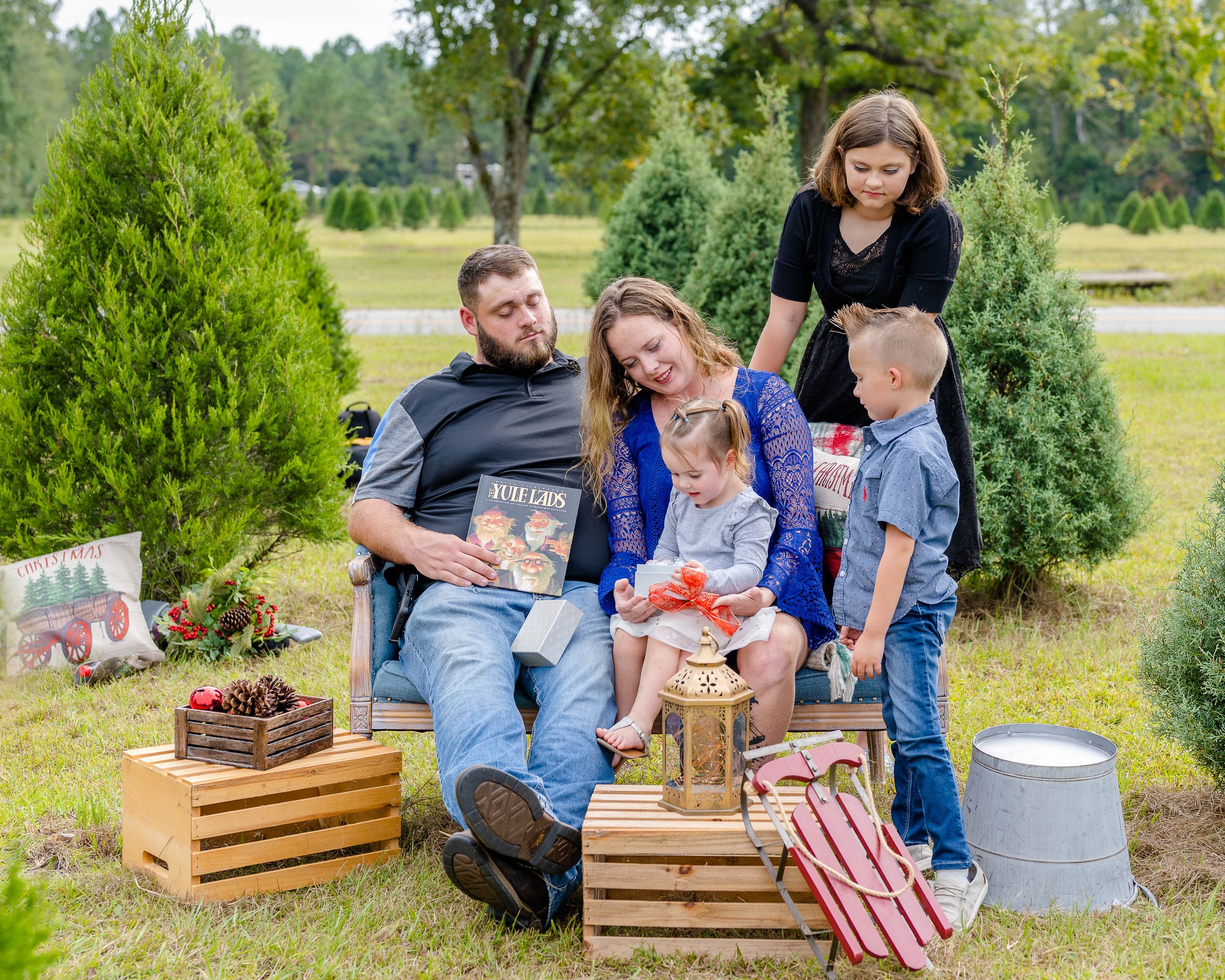 Family gathering at Christmas tree farm with wooden crates and holiday decorations among evergreens.