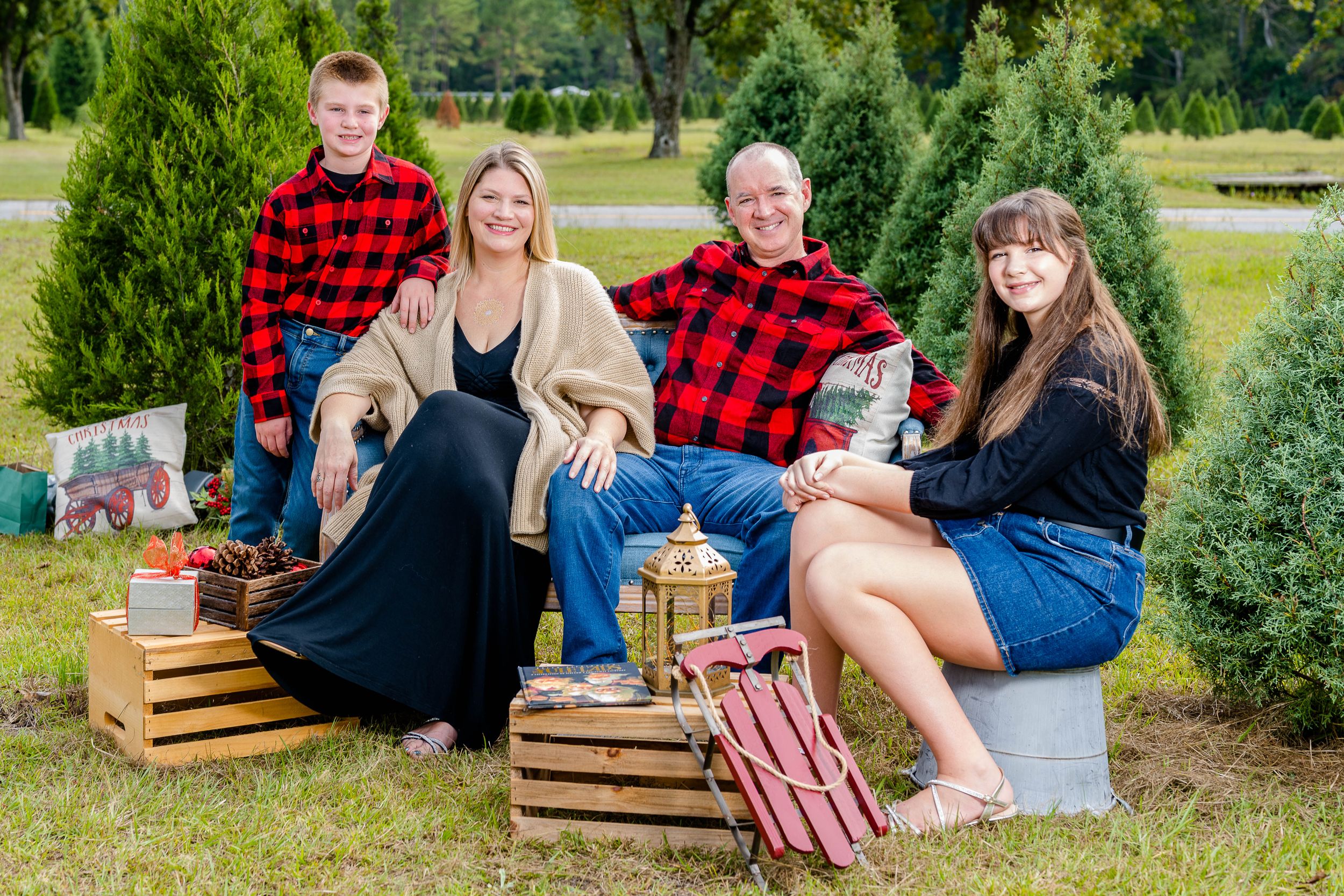Family group in buffalo plaid and casual wear poses at Christmas tree farm with wooden crate props.