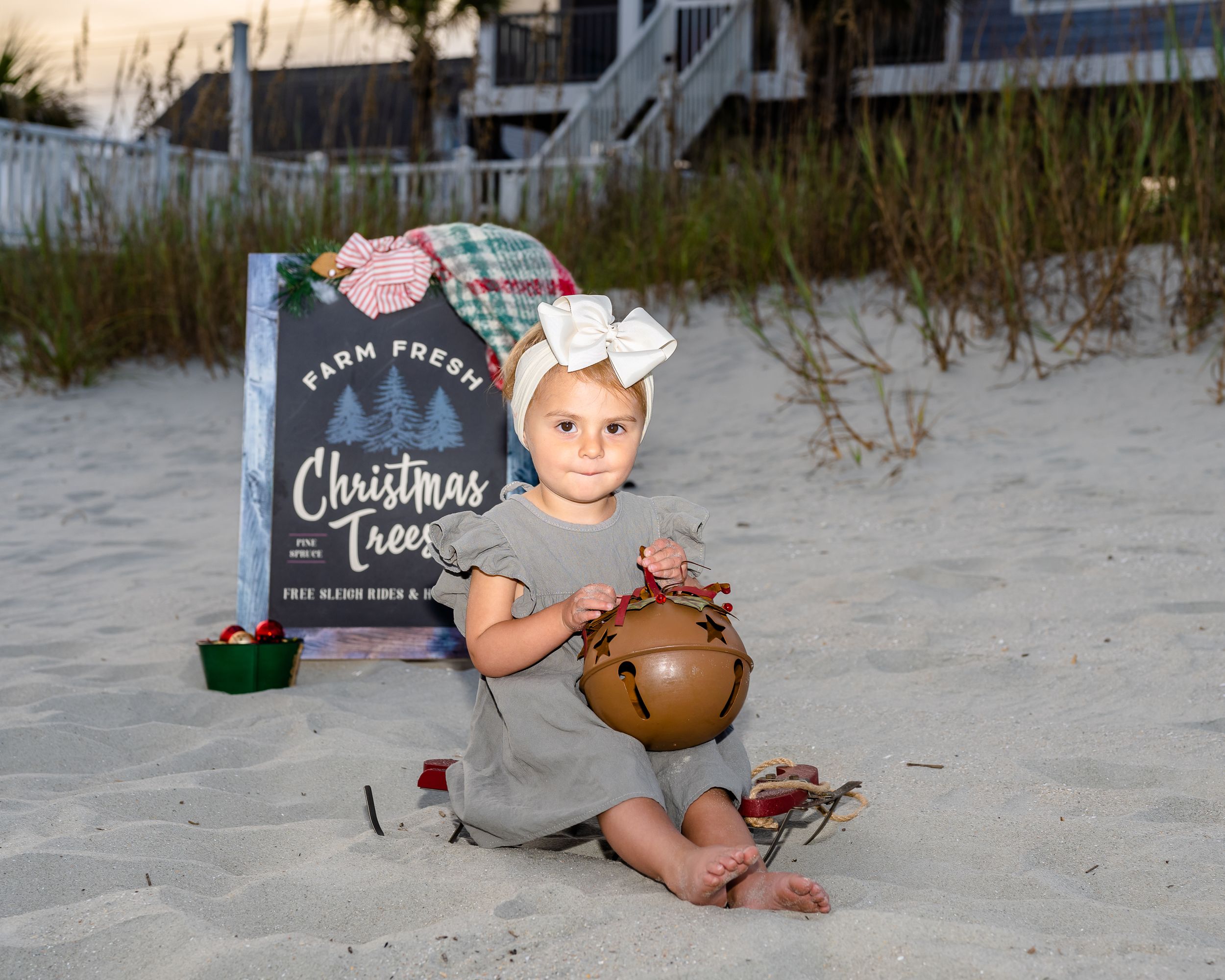 Small child sits on beach sand next to Christmas-themed book and decorative items at sunset.